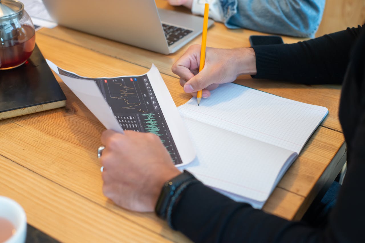 Close-up of hands writing notes on notebook during business meeting with laptop and charts.